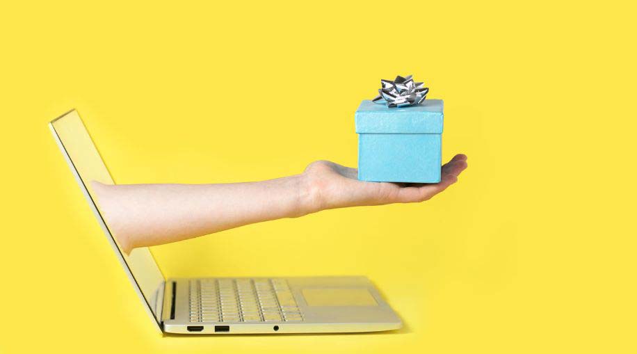 A hand holding a wrapped present coming out of a laptop screen against a yellow background.