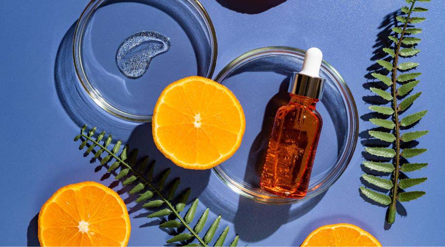 A bottle of vitamin C serum next to slices of oranges and a fern leaf against a dark blue background.