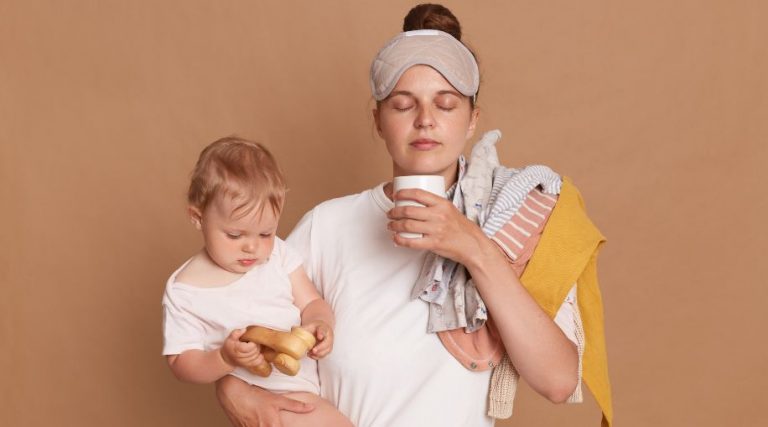 A new mom holding their baby while drinking coffee, looking tired.