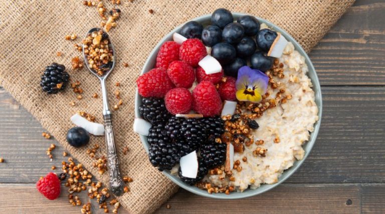 A bowl of oatmeal with different berries and almonds on top, sitting on a table beside a metal spoon.